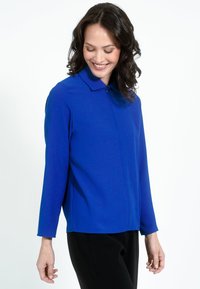 Királykék, hosszúujjú blúz, galléros nyakkal. Simább textúra és minimalista dizájn. Fekete nadrággal párosítva.