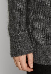 Ärmel eines grauen Strickpullovers mit einem gerippten Bündchen. Der Stoff zeigt ein strukturiertes Muster, das dunkle und helle Grautöne mischt.