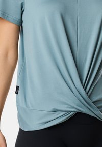 Ljusblå t-shirt i mjukt och elastiskt tyg, med en knuten design fram, korta ärmar och en liten märkesskylt vid fållen.