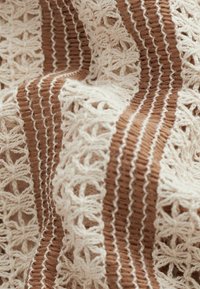 Tekstylia szydełkowe o naturalnym kremowym kolorze z naprzemiennymi brązowymi paskami. Wzór zawiera misternie wykonane geometryczne kształty i teksturowane wykończenie.
