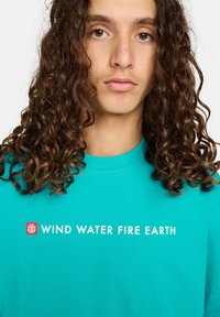 Young person with long curly brown hair wearing a turquoise shirt with text "WIND WATER FIRE EARTH" on front.