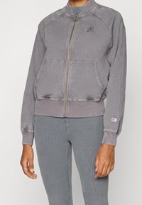Giacca bomber grigia con zip frontale, maniche raglan, due tasche frontali e un piccolo logo sul petto. Polsini e vita a costine.