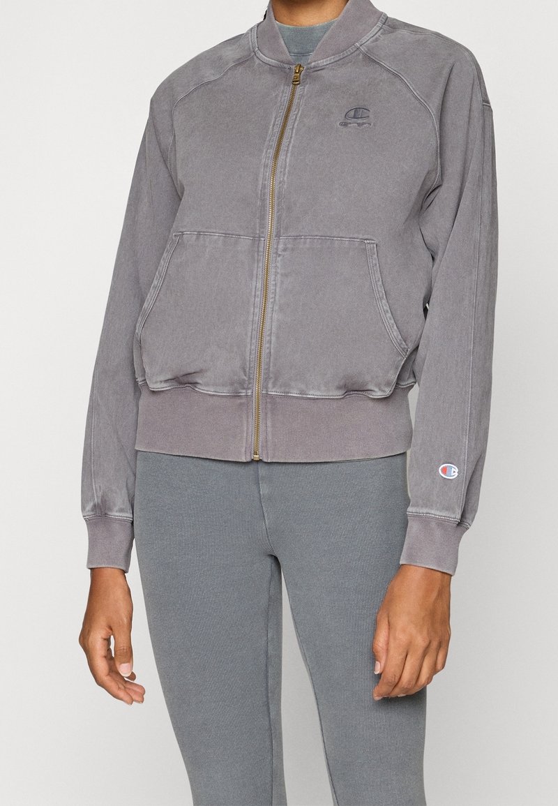 Giacca bomber grigia con zip frontale, maniche raglan, due tasche frontali e un piccolo logo sul petto. Polsini e vita a costine.