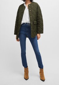 Olivengrön, vadderad jacka med tryckknappar, vit texturerad blus, mörkblå jeans och ljusbruna mocka ankellång stövlar med spetsig tå.