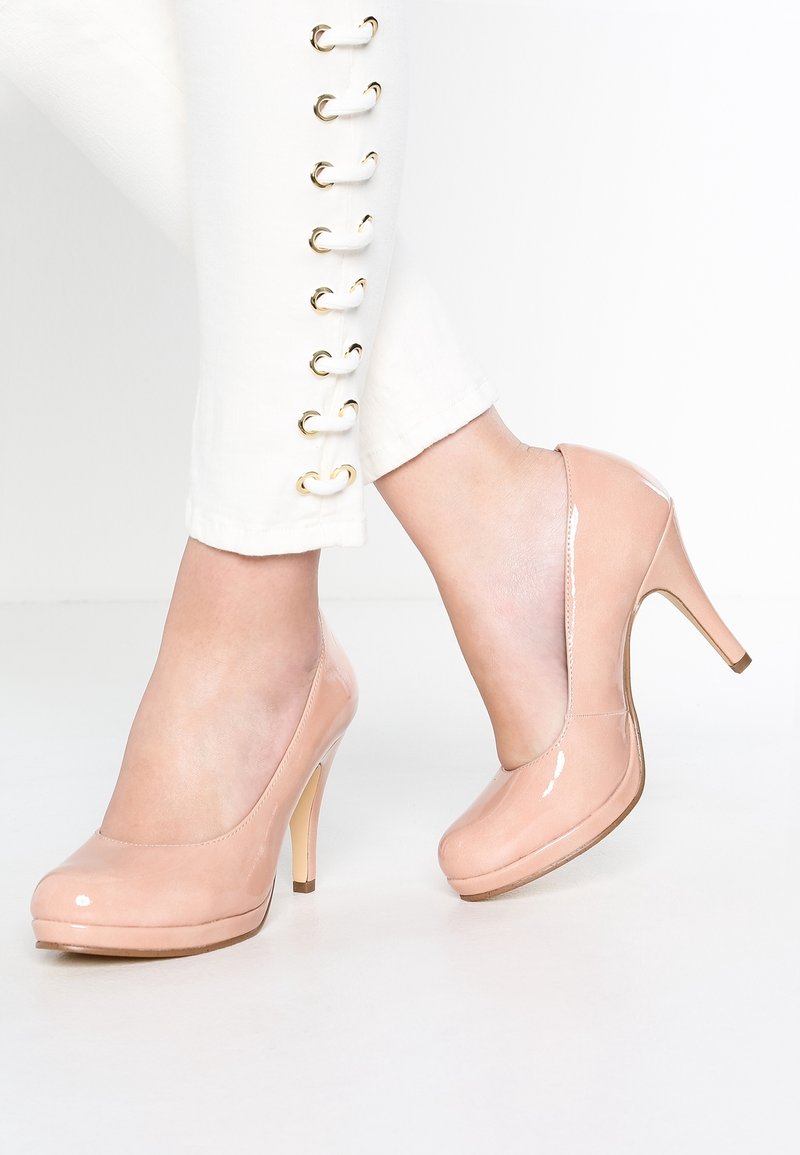 Femme portant un pantalon blanc avec des détails de boutons dorés et des chaussures à talons hauts en cuir verni nude, debout sur un sol blanc.