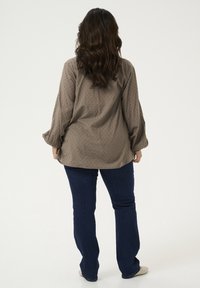 Khaki blouse met gestructureerde stippen, met lange mouwen en een losse pasvorm. Gecombineerd met donkerblauwe flared jeans en lichtgekleurde schoenen.