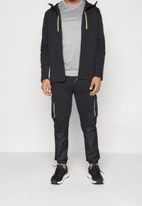 Zwarte jas met rits en groene accenten, gecombineerd met donkere broek met cargozakken. Model draagt zwarte sneakers. Grijs sportief T-shirt eronder.