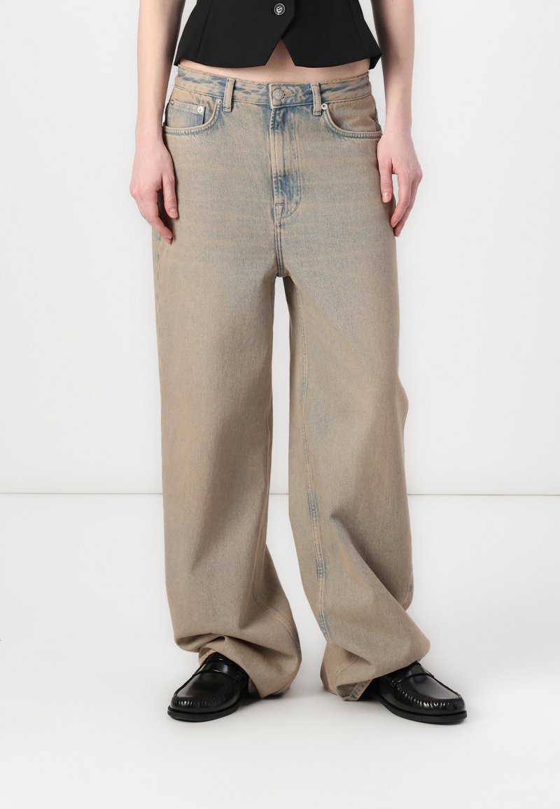 Jeans in denim beige chiaro a vestibilità rilassata, con una texture leggermente sbiadita, apertura della gamba ampia e design a cinque tasche. Indossati con scarpe nere.
