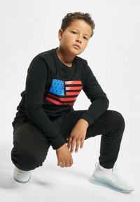 Zwarte lange mouwen shirt met een blauw en rood Amerikaans vlag graphic. Zwarte broek en witte sneakers met een doorzichtige zool.