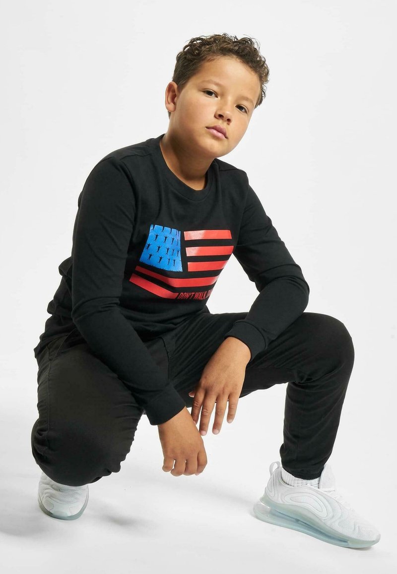 Zwarte lange mouwen shirt met een blauw en rood Amerikaans vlag graphic. Zwarte broek en witte sneakers met een doorzichtige zool.