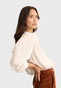 Blusa leggera color crema con maniche voluminose e dettagli di scollo arricciati, indossata con pantaloni in corduroy color ruggine.