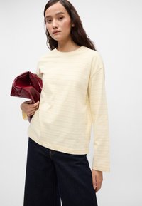 Ανοιχτό κίτρινο μακρυμάνικο πουκάμισο με ρίγες και στρογγυλή λαιμόκοψη, συνδυασμένο με σκούρα τζιν παντελόνια σε φαρδύ κόψιμο, και κρατώντας μια γυαλιστερή μπορντό τσάντα χειρός.