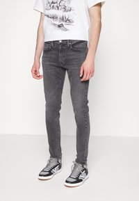 Slim fit, sötétszürke farmernadrág, klasszikus öt zsebes kialakítással. Fehér grafikai pólóval és fekete sportcipővel viselve.