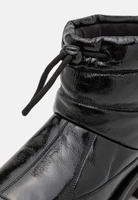 Black patent leather ankle boot featuring a padded top, textured surface, adjustable drawstring, and subtle stitching details.