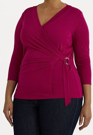 Kvinde iført en tætsiddende, magenta omslagsbluse med trekvartærmer og en sølvspændedetalje, kombineret med mørk denimjeans.