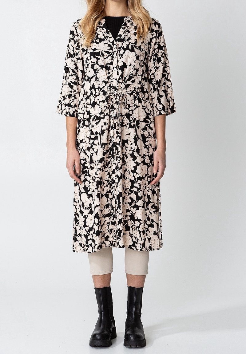 Femme portant une robe noire mi-longue à imprimé floral blanc, un pantalon beige court et des bottines noires, debout devant un fond uni.