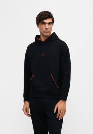 Jeune homme aux cheveux foncés portant un sweat à capuche noir avec un liseré orange et le mot HUGO sur la poitrine, debout devant un fond uni.