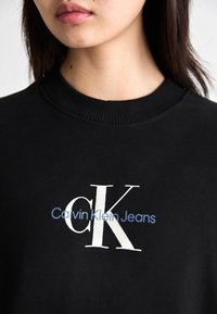 Črno bombažno pulover s ribanim ovratnikom, ki ima bele in modre napise "Calvin Klein Jeans" ter velik beli logotip "CK".