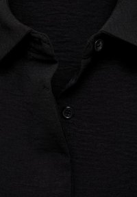 Chemise noire boutonnée avec un tissu texturé, dotée d'un col classique et d'un seul bouton noir au niveau du col. Design simple et épuré.