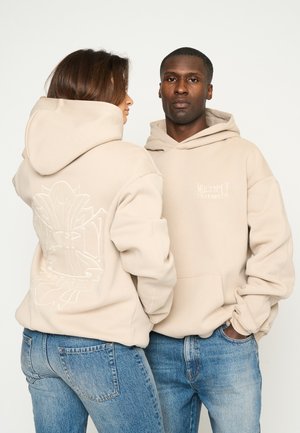 Sweatshirts beiges en matériau doux, avec une poche avant, une capuche à cordon et un motif floral brodé au dos. Portés avec un jean bleu.