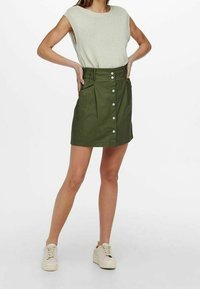 Grüne knielange skirt mit vorderem Knopfverschluss, geplätteten Taschen und einem taillierten Bund; kombiniert mit einem hellgrauen ärmellosen Oberteil und beigen Sneakers.