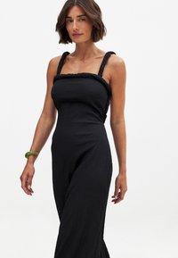 Svart jumpsuit med strukturerad tyg, tjocka flätade remmar, skräddarsydd överdel och utsvängda byxor. Ett grönt armband på vänster handled skapar kontrast.