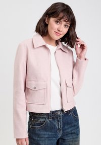 Giacca corta rosa realizzata in materiale morbido, con zip frontale, colletto a punta, due tasche anteriori e maniche lunghe, abbinata a jeans blu.