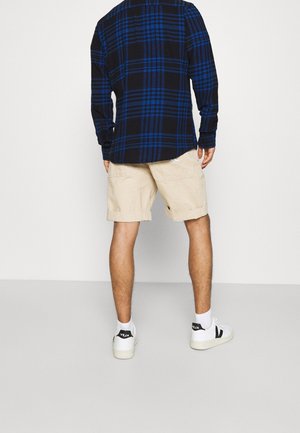 Homme portant un short beige, des baskets blanches, des chaussettes blanches et une chemise à carreaux bleue et noire à manches longues, de dos.