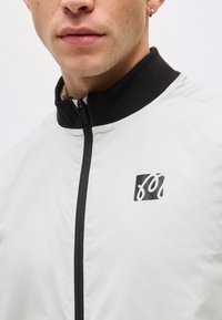 Gros plan sur une personne portant une veste blanche à fermeture éclair avec un col noir et un petit logo noir et blanc sur la poitrine.