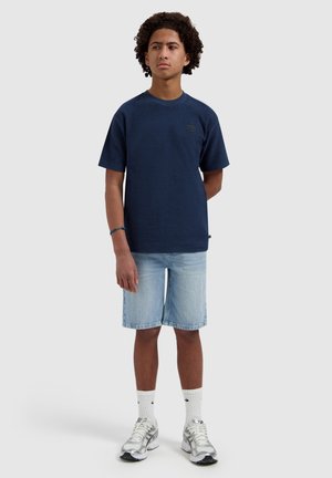 Teenager-Junge mit lockigem Haar, der ein dunkelblaues kurzärmeliges Hemd, helle Jeansshorts, weiße Nike-Socken und silberne Laufschuhe trägt und mit den Armen hinter dem Rücken steht.
