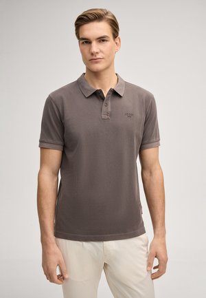 Junger Mann, der ein braunes kurzärmeliges Poloshirt und weiße Hosen trägt, steht vor einem einfarbigen hellen Hintergrund.