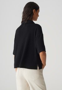 Pull noir à manches courtes avec un col, en tissu texturé, et fentes sur les côtés, associé à un pantalon blanc.