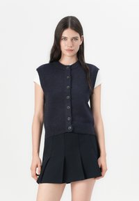Gilet navy in maglia con maniche corte, dotato di un collo alto, chiusura frontale con bottoni e orlo a coste, abbinato a una gonna plissettata nera.