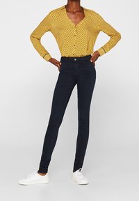 Blusa amarilla con estampado y detalles de botones, combinada con jeans ajustados oscuros y zapatillas blancas. Tela suave y diseño entallado.