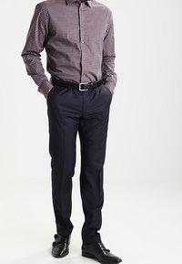 Camicia a maniche lunghe a scacchi in bordeaux e bianco, abbinata a pantaloni scuri blu navy. Presenta una cintura e scarpe nere lucide.