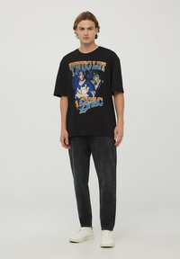 Czarny oversize t-shirt z graficznym nadrukiem Tupaca i napisem "THUG LIFE" w wyraźnym żółtym i niebieskim kolorze. Noszony z ciemnymi dżinsami i białymi trampkami.