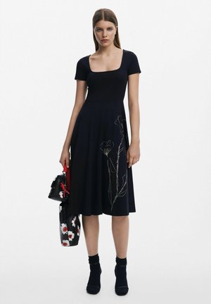 Abito navy con maniche corte, scollo quadrato e ricami floreali sul gonnellino. La modella tiene una borsa nera con dettagli rossi. Calze nere e tacchi.