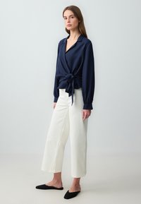 Blusa avvolgente blu navy con maniche lunghe e dettaglio a fiocco in vita, abbinata a pantaloni bianchi a gamba larga e scarpe nere slip-on.