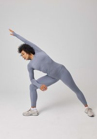 Grijze sportoutfit met lange mouwen en strakke leggings. De stof lijkt glad met een lichte glans. Witte sneakers maken de look af.