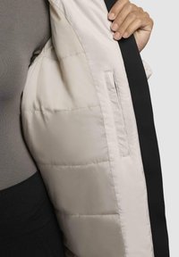 Puffer jacket with a light beige interior and black outer shell. Features a smooth texture, side pocket, and quilted stitching.