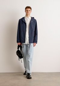 Navy lightweight jacket with hood, white shirt, light blue relaxed-fit trousers, and black shoes; holding a small black bag with front stripes.