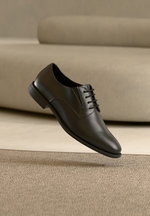 Sort læder Oxford-formel sko med snørebånd, placeret i luften over et beige tæppe i et minimalistisk rum.