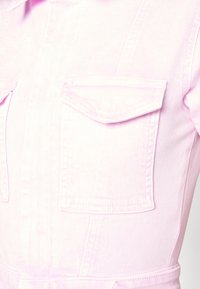 Ljusrosa denimoverall med två bröstfickor, synlig sömnad och en figurnära design med bred midjeresår. Slät struktur.