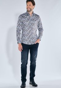 Langärmliges Hemd mit floralen Mustern in Blau und Weiß, aus leichtem Material. Kombiniert mit dunkelblauen Jeans und schwarzen Sneakers.