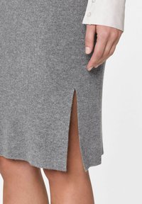 Hellgrauer gerippter Strickrock mit seitlichem Schlitz, der eine weiche Textur und einen leicht unregelmäßigen Saum aufweist. Eine Hand greift nach dem Rockdetail.