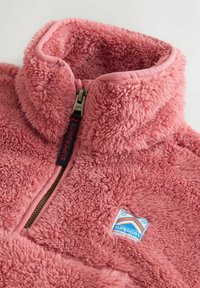 Lyserød fluffy fleecejakke med en lynlås foran og et lille mærkebadge med teksten "Superdry Mountain Sport" på brystet.