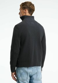 Dunkelgrauer, gerippter Strickpullover mit hohem Kragen, der eine einheitliche Textur und subtile Rückendetails aufweist. Kombiniert mit hellblauen Jeans.