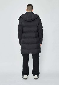 Manteau matelassé noir avec une capuche, présentant un design matelassé et une silhouette longue. Porté par-dessus un pantalon cargo noir et des baskets blanches.