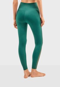 Leggings verts à taille haute, fabriqués en matériau extensible. Présente des panneaux latéraux transparents et une finition lisse et mate.