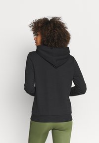 Černá mikina s měkkou texturou, má velkou kapuci a žebrovaný lem. Nosí se s zelenými legínami, což ukazuje uvolněný střih a jednoduchý design.